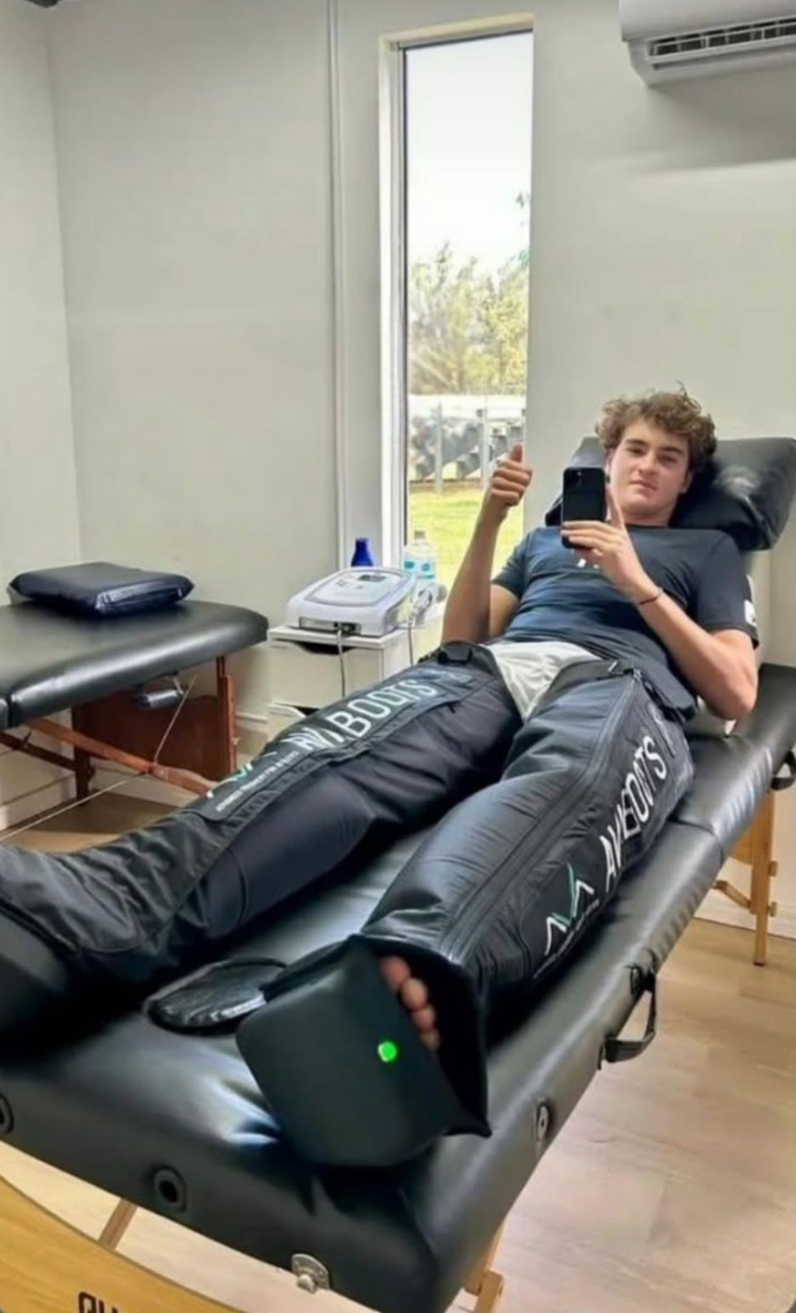 Tennis player João Fonseca lying on a treatment table in a clinic, taking a selfie with his phone and giving a thumbs up, with both legs inside the AVABoots compression boots.