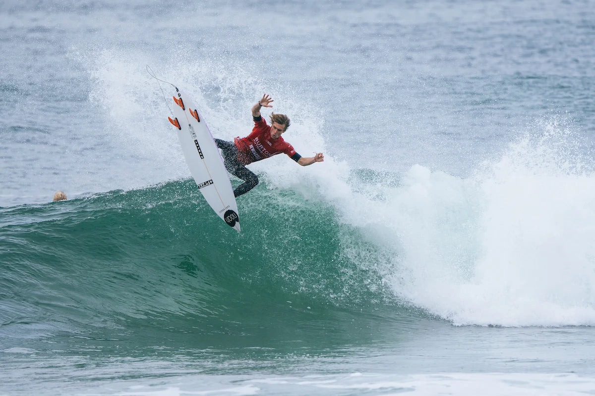 Surfista executa uma manobra aérea sobre uma onda, com a prancha levantada e o corpo em posição de equilíbrio. Usa fato de surf preto e licra vermelha de competição, com o mar a formar espuma ao fundo.