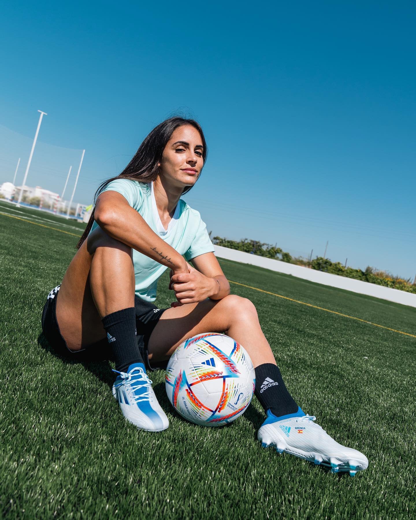 Jogadora de futebol sentada no relvado de um campo desportivo, com expressão confiante. Veste equipamento desportivo leve, com chuteiras brancas e azuis da Adidas, e tem uma bola oficial de futebol junto aos pés sob um céu limpo e azul