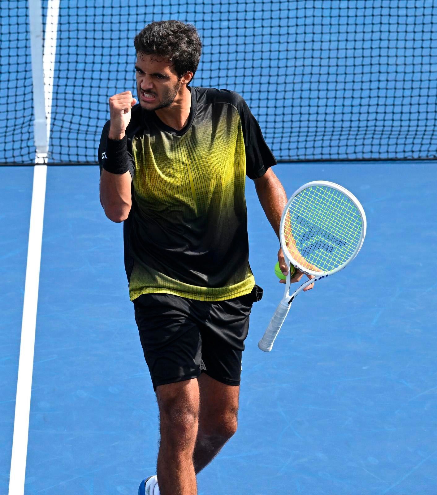 Tennis player celebrates with a clenched fist and a determined expression, standing on a blue hard court. He is wearing a black outfit with yellow details and holding the racket in his left hand.