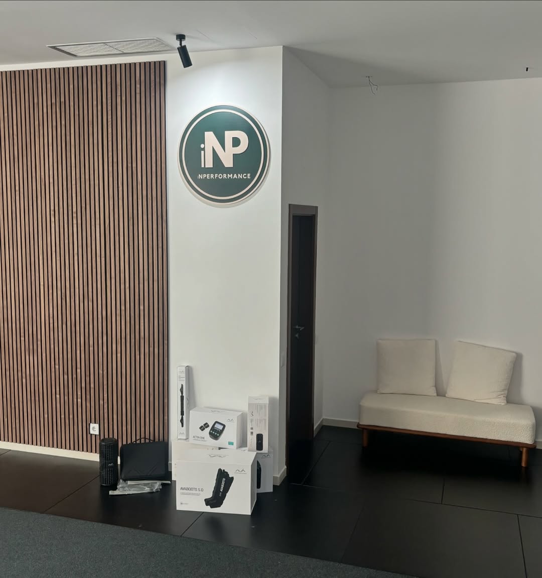 Espacio interior moderno con el logotipo circular de iNPERFORMANCE en la pared. En la esquina, se exhiben varios productos de recuperación de la marca AVA, incluidas cajas de botas de compresión, un rodillo de masaje y otros accesorios. A un lado, un sofá blanco de estilo minimalista.