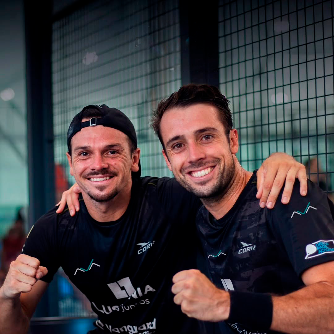Two padel players celebrate with joyful expressions and clenched fists, embracing next to a metal net.
They are wearing black shirts with sponsor logos and the Cork brand, in an indoor competition setting.