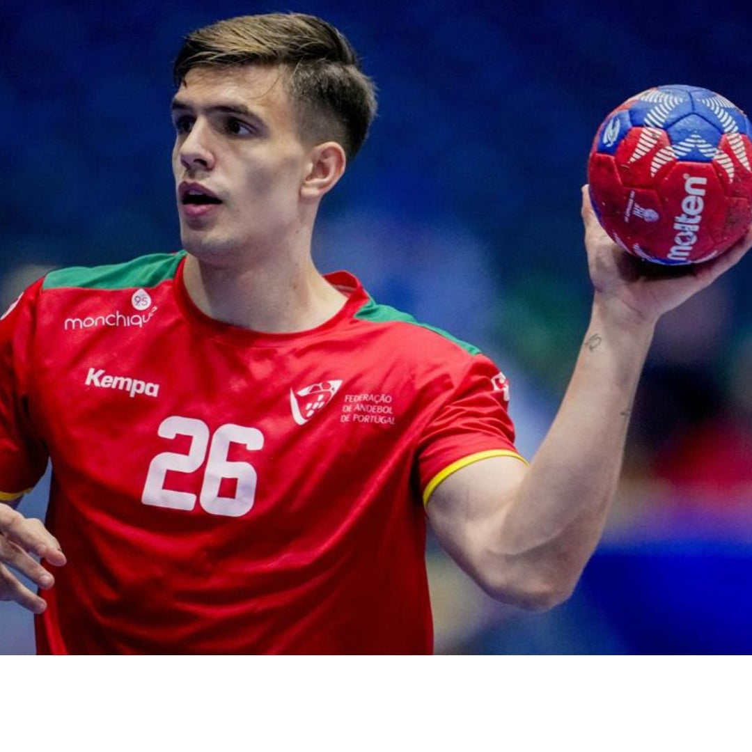 Player of the Portuguese national handball team, wearing number 26, holds the ball with his right hand and prepares to make a pass or a shot, dressed in the official red kit with green and yellow details.