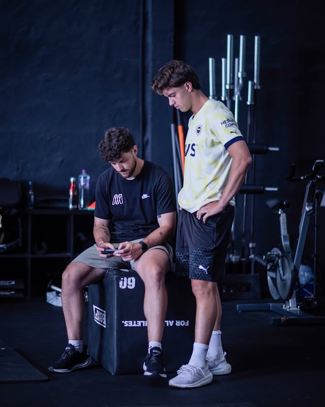 Two men in a gym. One is sitting on a plyometric box using a phone, wearing a black t-shirt with the “Move Better” logo. The other stands beside him, watching the screen, dressed in yellow and black sportswear.