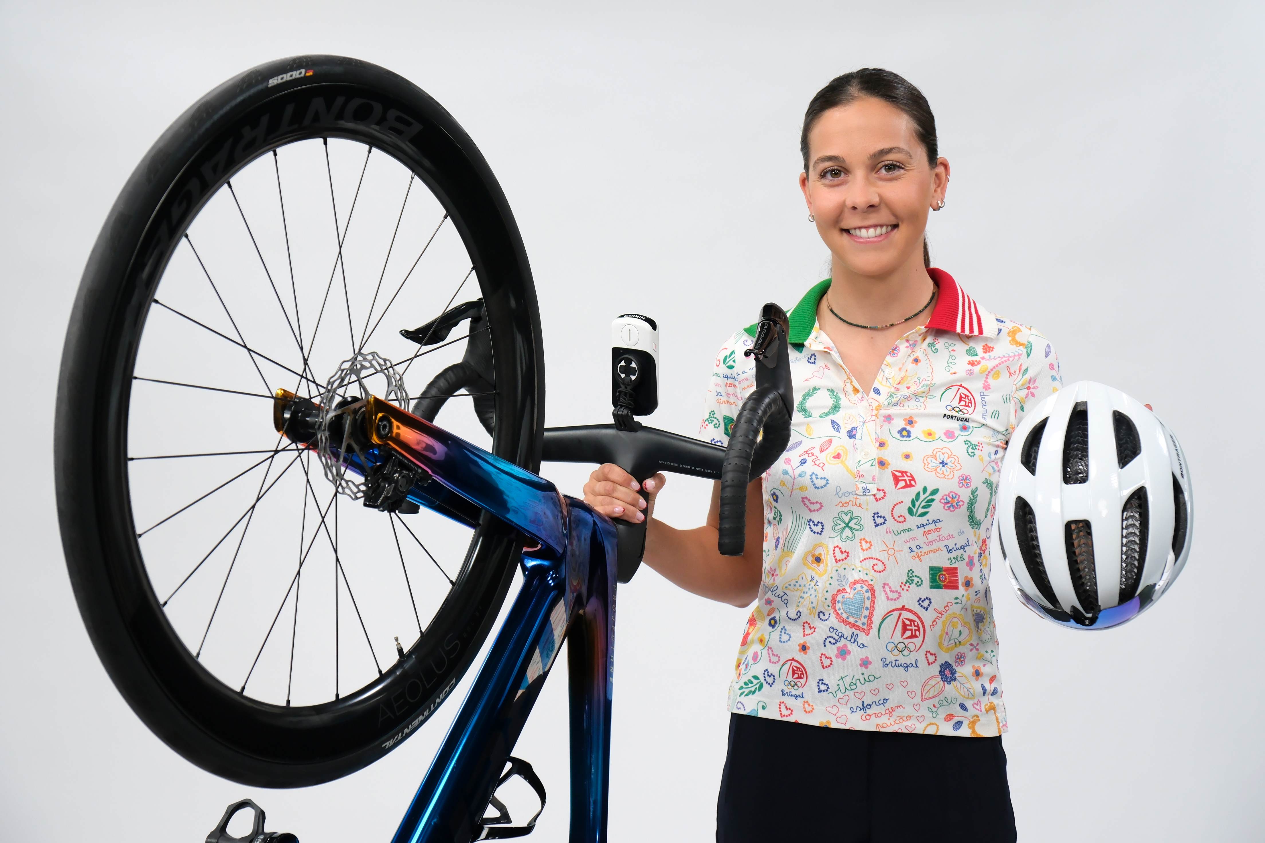 Ciclista portuguesa sorri enquanto segura uma bicicleta de estrada com uma mão e um capacete branco com a outra. Veste uma camisola colorida com símbolos e palavras desenhadas, incluindo a bandeira de Portugal e o logótipo do Comité Olímpico de Portugal.