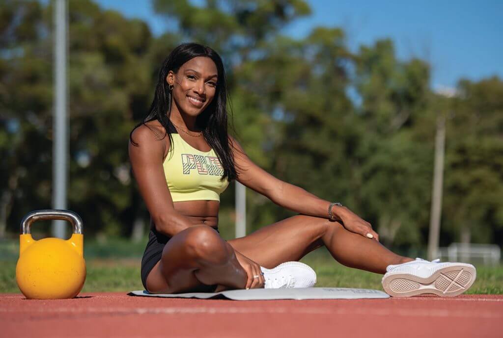 Athlete sitting on an outdoor training mat, smiling, with a yellow kettlebell beside her.
She is wearing a yellow sports top and black shorts, in an athletics track setting with trees in the background.