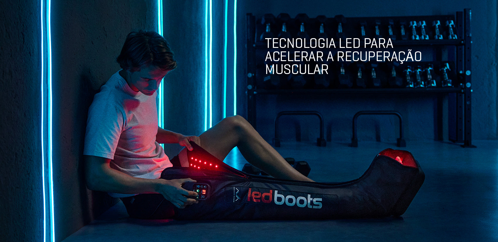 Atleta sentado num ginásio escuro com iluminação azul, utilizando um dispositivo de compressão com LEDs vermelhos nas pernas, com o texto “Tecnologia LED para acelerar a recuperação muscular” visível no canto inferior.