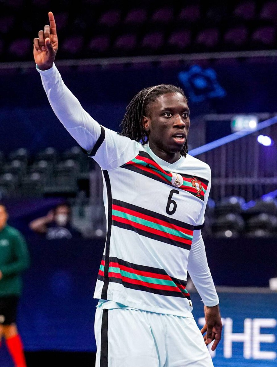 Jugador de la selección portuguesa de fútbol sala viste el uniforme alternativo blanco con franjas verde, roja y negra, celebrando con el brazo derecho levantado y el dedo índice apuntando hacia arriba.
El número 6 es visible en la camiseta, así como el emblema de campeón mundial de la FIFA.