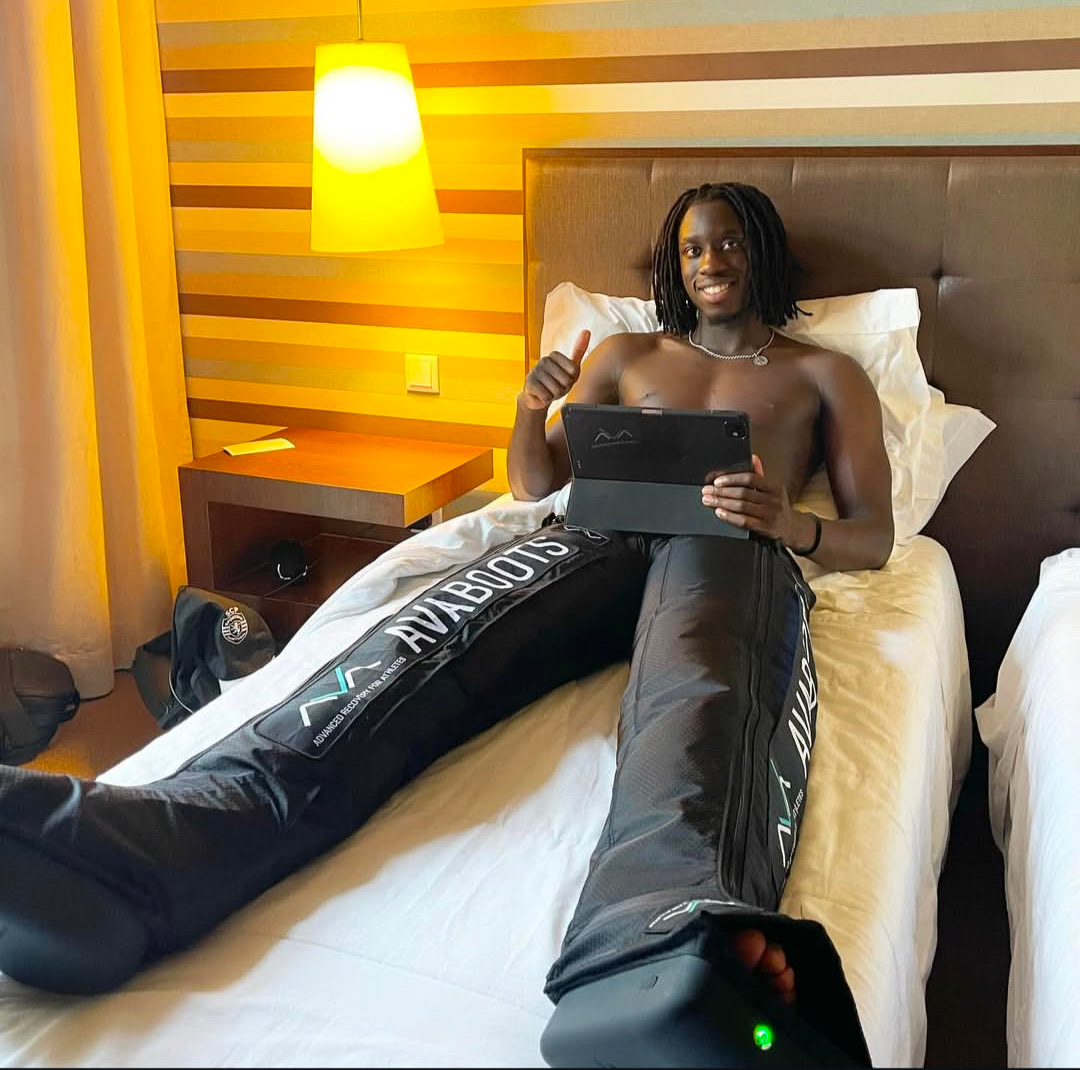 Atleta deitado numa cama de hotel a utilizar botas de compressão AVABOOTS para recuperação muscular, enquanto segura um tablet e faz sinal positivo com o polegar.