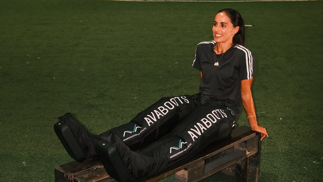 Athlete Brenda Perez sitting on a wooden bench on artificial turf, smiling, with both legs inside the AVABoots compression boots during a nighttime recovery session on a sports field.