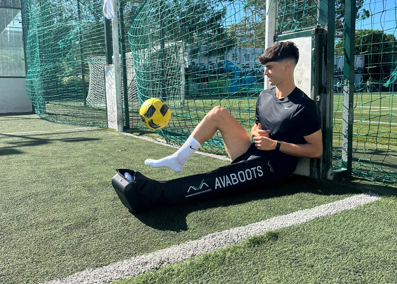 Footballer Rodrigo Ribeiro sitting against a goalpost on an artificial turf pitch, smiling, wearing an AVABoots compression boot on his right leg as a ball flies through the air.