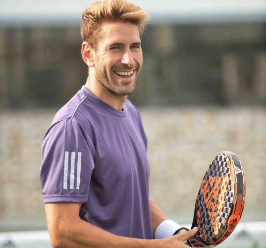 Jugador de pádel sonríe a la cámara mientras sostiene una pala con diseño naranja y negro.
Viste una camiseta morada con rayas blancas en la manga y una muñequera blanca en la muñeca izquierda, en un entorno deportivo al aire libre.