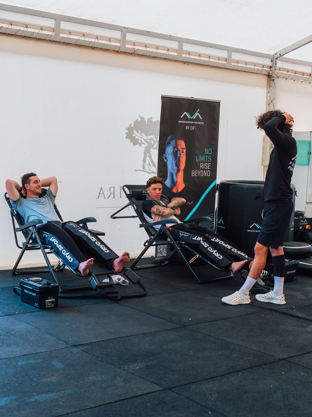 Três atletas em momento de recuperação muscular numa zona de treino interior. Dois deles estão deitados em cadeiras reclináveis com botas de compressão "Cryo Sport" nas pernas, enquanto o terceiro está de pé. Ao fundo, um cartaz com a imagem de Cristiano Ronaldo e o slogan “No Limits Rise Beyond”.