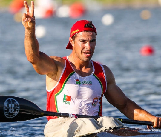 Portuguese canoeist celebrates inside the kayak, raising two fingers in a victory sign.
He wears a red cap turned backwards and a competition outfit in the colors of Portugal with official logos, holding the paddle with his right hand over the water.