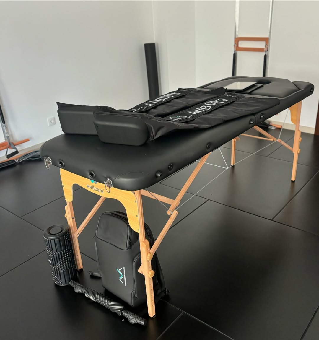 Mesa de massagem profissional com botas de compressão AVABOOTS dispostas por cima. No chão, ao lado da mesa, estão um rolo de libertação miofascial, uma mochila AVA e um stick de massagem, num espaço moderno de fisioterapia ou recuperação.