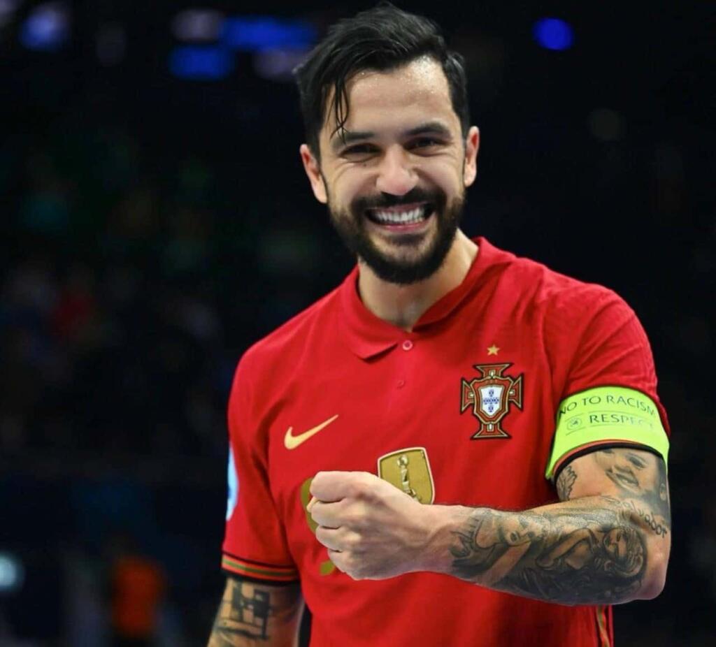 Jugador de la selección portuguesa de fútbol sala sonríe ampliamente mientras muestra su brazo tatuado.
Viste el uniforme oficial rojo con el emblema de campeón y el brazalete de capitán con la inscripción “No to racism – Respect”.